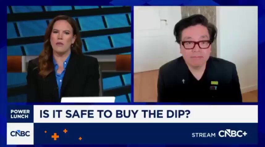 The image shows a news segment featuring two people, one woman wearing glasses and the other man dressed in a suit. They are both talking about Bitcoin and cryptocurrency, with the woman stating that Bitcoin is safe to buy the Dip. The man responds by saying that Bitcoin is not safe to buy the Dip because it's a speculative asset and its value can be highly volatile.