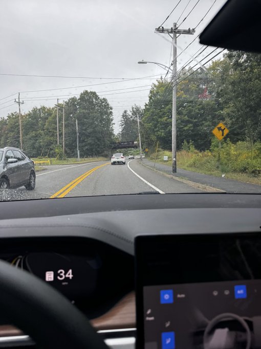 A view from inside a Tesla showing the dashboard displaying a speed of 34 mph, with a rain-speckled windshield revealing a road, power lines, trees, and a yellow traffic sign ahead.
