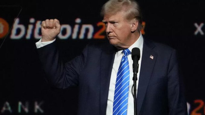 Former U.S. President Donald Trump stands at a podium with a clenched fist, in front of a backdrop displaying "Bitcoin 2024" and partially visible "x" and "BANK" text.