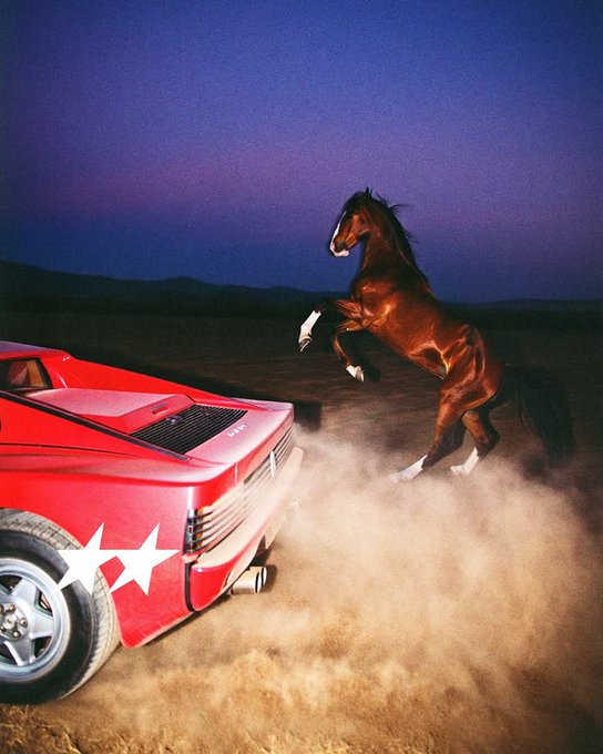 An artistic photograph of a rearing brown horse next to a red sports car kicking up dust against a purple twilight sky.