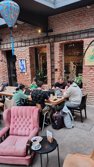 A group of people sit around a wooden table working on laptops in a brick-walled cafe courtyard, with a decorative blue and orange paper lantern hanging above and a pink tufted armchair in the foreground beside a small table holding a coffee cup.