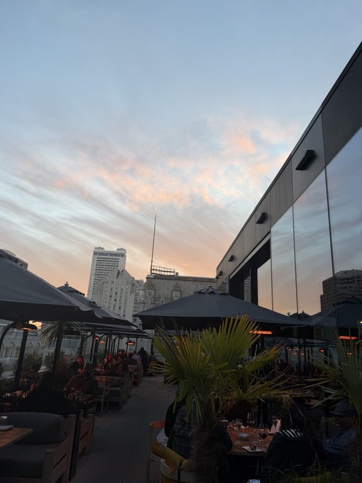 A rooftop terrace scene at dusk features black umbrellas, palm plants, and people seated at tables, with a sunset sky and city skyscrapers in the background.