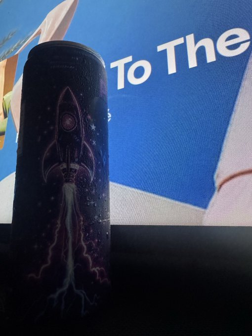 A black cylindrical object, possibly a light stick or bottle, decorated with a rocket design sits in front of a screen displaying the text 'To The'.