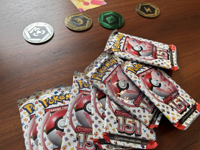 Multiple Pokemon Trading Card Game booster packs from the Scarlet & Violet 151 set scattered on a wooden table alongside several colored game tokens or coins.