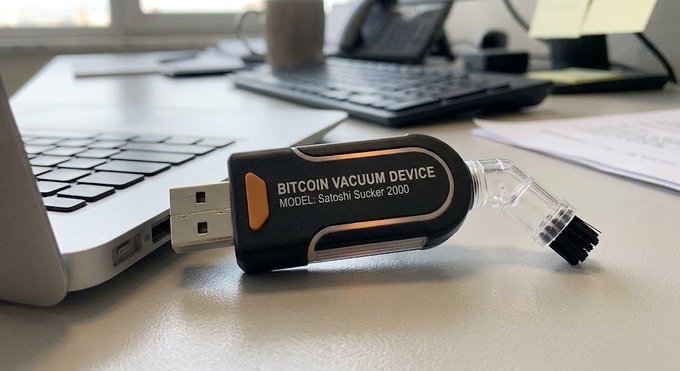A novelty USB device labeled 'BITCOIN VACUUM DEVICE - MODEL: Satoshi Sucker 2000' sits plugged into a laptop on a desk, featuring a clear glass attachment at the end.