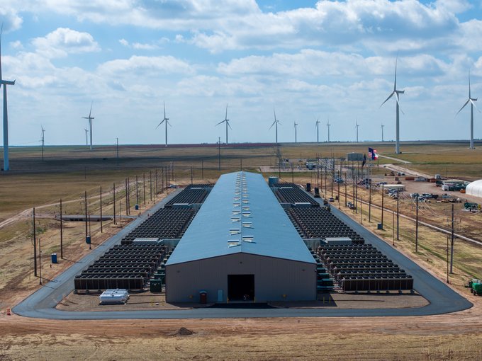 Aerial view of a large industrial facility with rows of black containers flanking a central warehouse building, set in an open plain with numerous wind turbines on the horizon and a Texas state flag flying nearby.