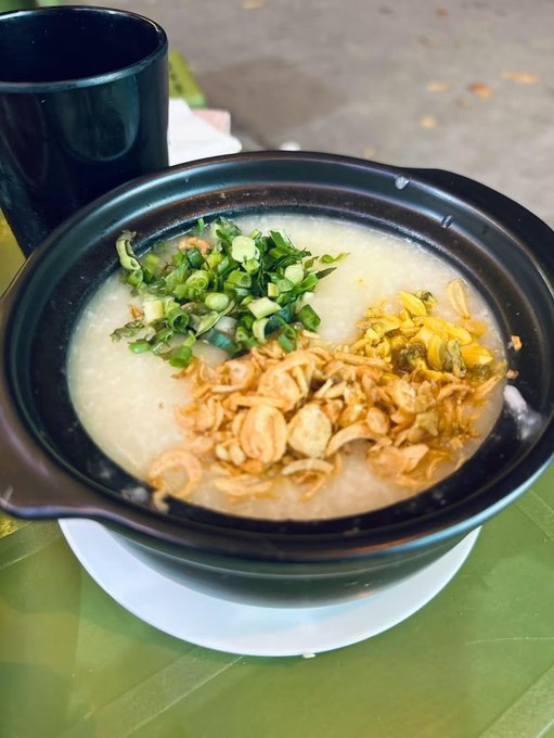 A bowl of congee or rice porridge topped with crispy fried shallots, chopped green onions, and pickled vegetables sits on a white plate.