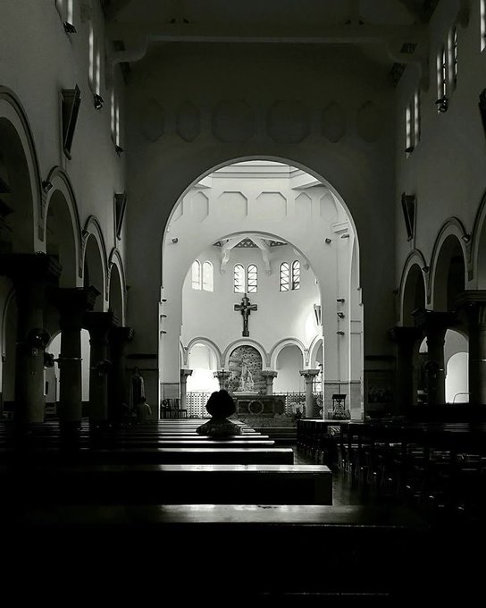 A high-contrast black and white photograph looking down the aisle of a church toward an illuminated altar with a cross, framed by arches.