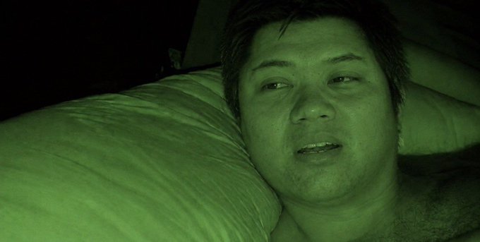 A close-up selfie-style photo of a man resting his head on a white pillow in dim, greenish lighting.