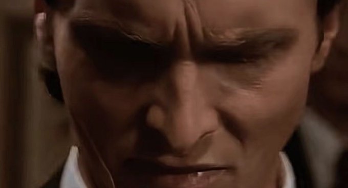 A close-up still of a man with a stern expression, his eyes cast downward, wearing a dark suit with a white collar.