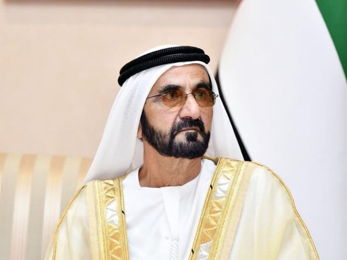 The image features a man wearing traditional Arab clothing, including a white robe with gold trimming. He is seated in front of a flag that displays the colors green, white, and red. The man appears to be looking off into the distance or deep in thought. In the background, there are two chairs visible, one closer to the foreground and another further back.