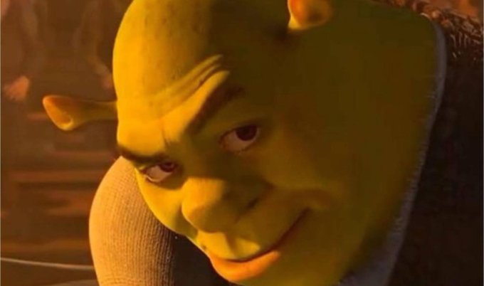 A close-up CGI rendering of Shrek's face with a sly, knowing expression in warm orange-toned lighting.