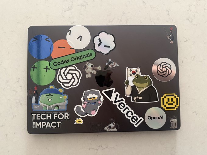 A black laptop covered in various stickers, including ones for 'Codex Originals', 'TECH FOR IMPACT', 'vercel', 'OpenAI', Vercel's triangle logo, and several cartoon characters.
