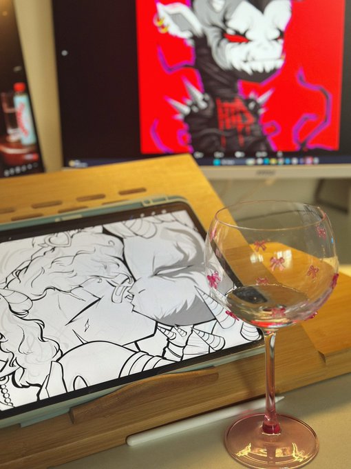 Workspace setup featuring a wine glass with decorative pink flowers in the foreground, with a tablet displaying black and white artwork and a monitor showing a red-tinted character illustration in the background.