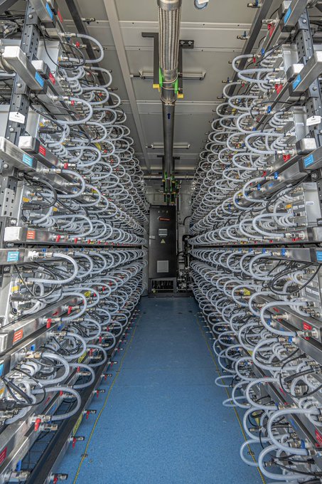 A data center aisle lined with dense cryptocurrency-mining hardware, cooling cables, and racks, illustrating the hosted mining infrastructure discussed in the post. The image itself has no prominent readable names, tickers, or numbers beyond small equipment labels.