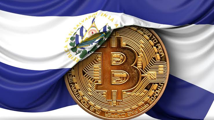 The El Salvador flag with its coat of arms draped behind a large golden Bitcoin coin featuring circuit-like patterns.