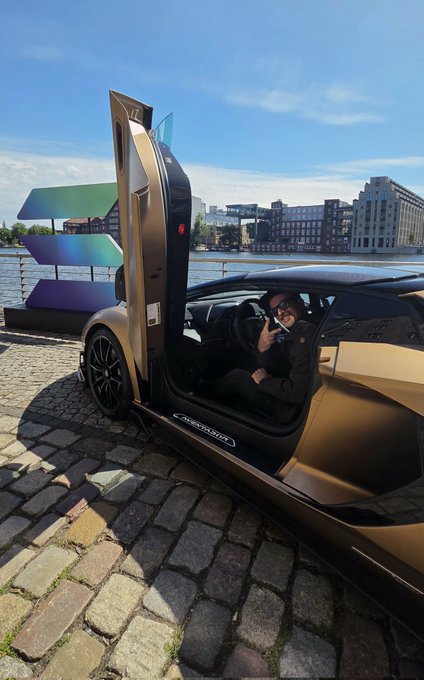 The image features a person sitting inside a car, with their head out of the open door. The car appears to be a sports car or a luxury vehicle, as indicated by its design and appearance. The car's position next to a brick walkway suggests that it might be parked in an urban setting. In the background, there are buildings visible, indicating that this scene takes place in a city environment. The image also contains text elements such as "GM" and "a great start to week #25 Still lost in Berlin - and I love it See us later at Ping Pong Mixer & USDC Splits".