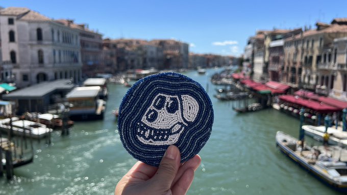The image features a hand holding up a blue and white coaster with a skull design, which appears to be made of beads or embroidery. The coaster is located near a body of water, possibly a canal, as there are several boats visible in the background. The caption on the image reads "GM from Venice! I’m actually not here but these beautiful photos are shared by our fellow ape @elethera_art @siw0bbyte @BoredApeYC What's cool & what makes me happy is that they have carried this coaster".