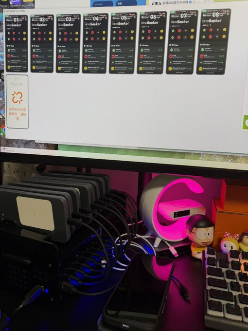 A monitor displays the JUST.FOTO app showing eight preview screens of a 'Seeker' design, with multiple smartphones connected via cables to charging stations below alongside headphones and figurines.