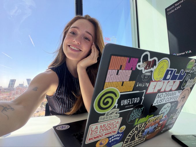 A smiling woman with long hair sits at a desk in front of a window, her laptop covered in stickers including 'JOOPBLAP', 'BLU', 'EPICO', 'NO DRAMA JUST COFFEE', 'LA MUSICA ES DROGA' and 'Smashing Corner'.