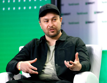 A man wearing a black cap and black overshirt speaking while gesturing with both hands, seated against a green branded backdrop.