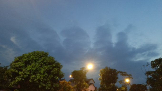 A dusk or dawn sky features scattered clouds, with silhouettes of trees and rooftops illuminated by glowing street lamps below.