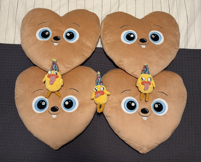 Four heart-shaped brown plush pillows with big cartoon eyes, each accompanied by a small yellow duck plush wearing a colorful party hat, arranged on a dark surface.