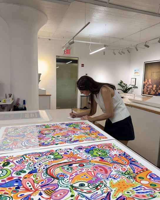 A woman in a white top and black skirt stands at a large table in a brightly lit gallery space, working on or examining vibrant, colorful psychedelic artwork featuring cartoon faces and abstract patterns.