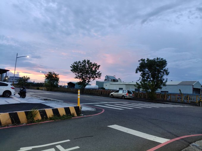 An outdoor street scene at sunset with pink and blue clouds, showing a road intersection, trees, buildings, and a car passing by.
