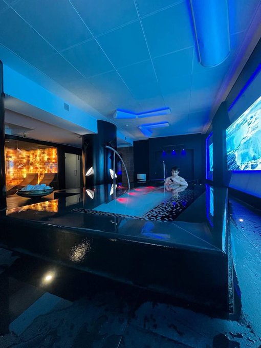 A dimly lit, luxurious indoor pool and spa area is illuminated by vibrant blue lighting, featuring a person sitting in the bubbling hot tub near a lit wall display.