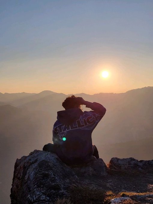 A person wearing a Metallica hoodie sits on a rocky cliff edge at sunrise, shielding their eyes while gazing out over misty mountain ranges.