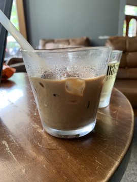 A close-up of an iced coffee drink with a straw in a clear glass sitting on a wooden table, with a second cup partially visible.