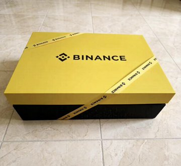 The image features a yellow box with a black ribbon, sitting on a tiled floor. The box has "Binance" written on it in white letters. The text also includes the words "Binance", "binance", "BINANCE", and "BINANCE". This suggests that the box is likely related to Bitcoin or another cryptocurrency, possibly as a gift or promotional item for the Binance cryptocurrency exchange platform.