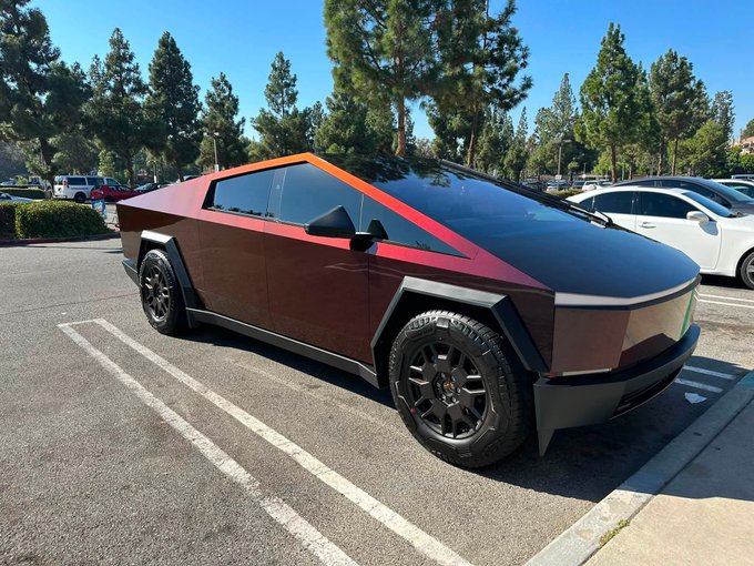 A red Tesla Cybertruck "CyberBeast" is parked in a shopping center lot, matching the owner's introduction of their new vehicle. The distinctive angular design and dark wheels of the electric pickup are clearly visible in the sunny outdoor setting.