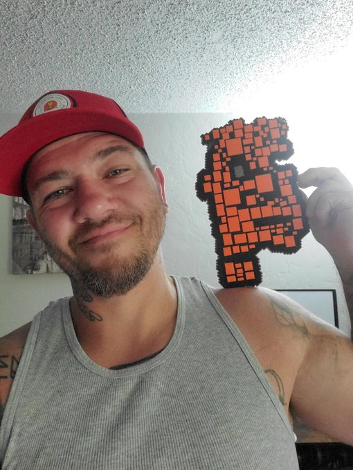 A bearded man in a red cap and gray tank top smiles while holding up a pixel-art-style red and black crocheted figure.