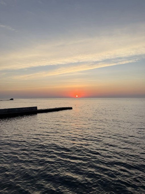 A calm seascape at sunset with the orange sun low on the horizon, a concrete pier extending into the water on the left, and soft pastel clouds above.