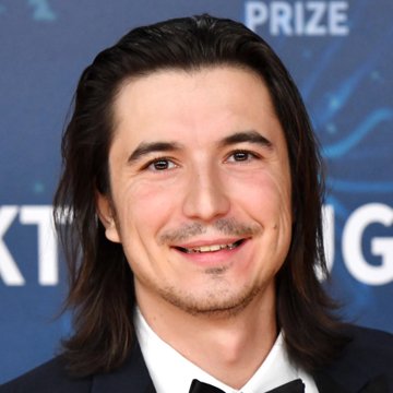 A man with long dark hair wearing a tuxedo smiles at what appears to be a formal event, with partial text "PRIZE" and "G" visible on the backdrop behind him. The image doesn't directly relate to the news about Robinhood Chain's Total Value Locked surpassing $600 million, suggesting the person pictured may simply be associated with the announcement in an unrelated context.