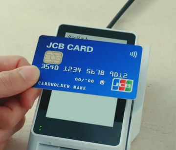 A hand holds a blue JCB Card (ending in 9012) near a payment terminal, visually representing the traditional card payment method that JCB aims to supplement with new USDC stablecoin payment options for foreign visitors to Japan.
