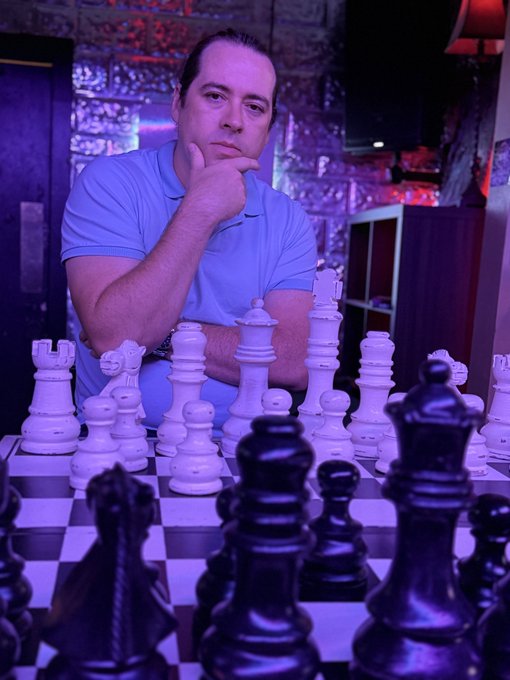 A man in a light blue polo shirt poses thoughtfully with his hand on his chin behind a large chess set, with a dark piece prominently in the foreground.