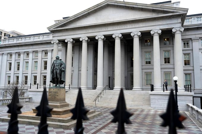 The United States Treasury Department building in Washington D.C., featuring its iconic neoclassical columns and a statue in the foreground, viewed through a black iron fence.