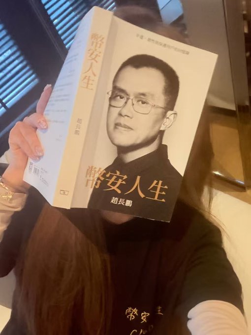 A woman holds up a Chinese-language book titled "幣安人生" (Binance Life) featuring a portrait of CZ (赵长鹏), the Binance founder, on the cover, visually reinforcing the post's theme about CZ's appearance in Hong Kong and his connection to the $Lin meme token.