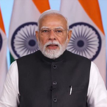 A man with a white beard and glasses, wearing a black vest over a white shirt, stands in front of two Indian national flags, likely making an official statement. The image is associated with news of India's central bank (RBI) supporting a nationwide ban on cryptocurrency.