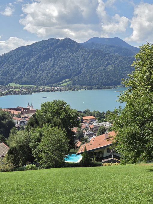 A picturesque view of Tegernsee Lake in Bavaria, Germany, with turquoise waters nestled between lush green mountains, overlooking a charming village with red-roofed homes and a swimming pool in the foreground. The scenic alpine landscape perfectly captures the serene "Good Morning CT" vibe from the elevated vantage point.