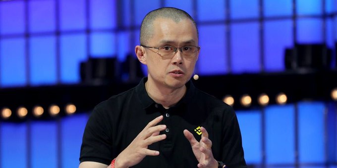 A man in a black polo shirt and glasses speaks at a panel, gesturing with both hands against a backdrop of blue illuminated panels.