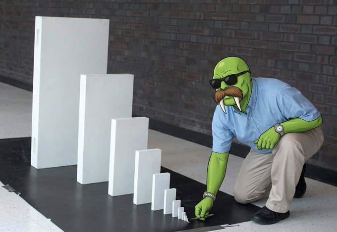 A person wearing a blue shirt and khaki pants poses as a green orc-like figure examining white rectangular blocks arranged in decreasing size.
