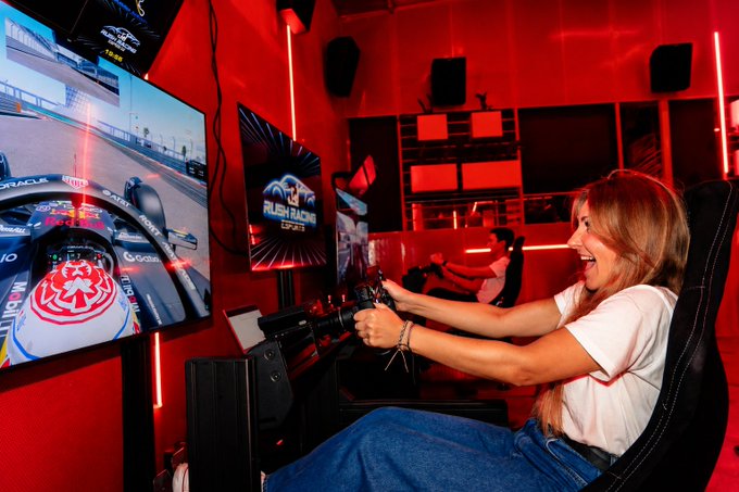 A woman in a white t-shirt and jeans excitedly grips a racing wheel controller while playing a Formula 1 racing simulator in a red-lit gaming room, with an Oracle Red Bull Racing car visible on the curved monitor.