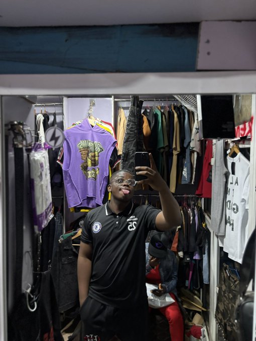 A young man taking a mirror selfie with his phone while sticking his tongue out, standing in what appears to be a clothing store or closet with various garments hanging behind him.