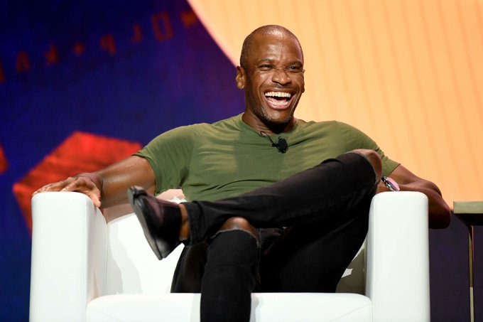The image features a man sitting in a chair with his legs crossed, smiling at the camera. The background of the photo shows a blue wall with an orange border, which appears to be part of a stage or presentation setting. There are also chairs visible in the scene, one near the foreground and another further back.