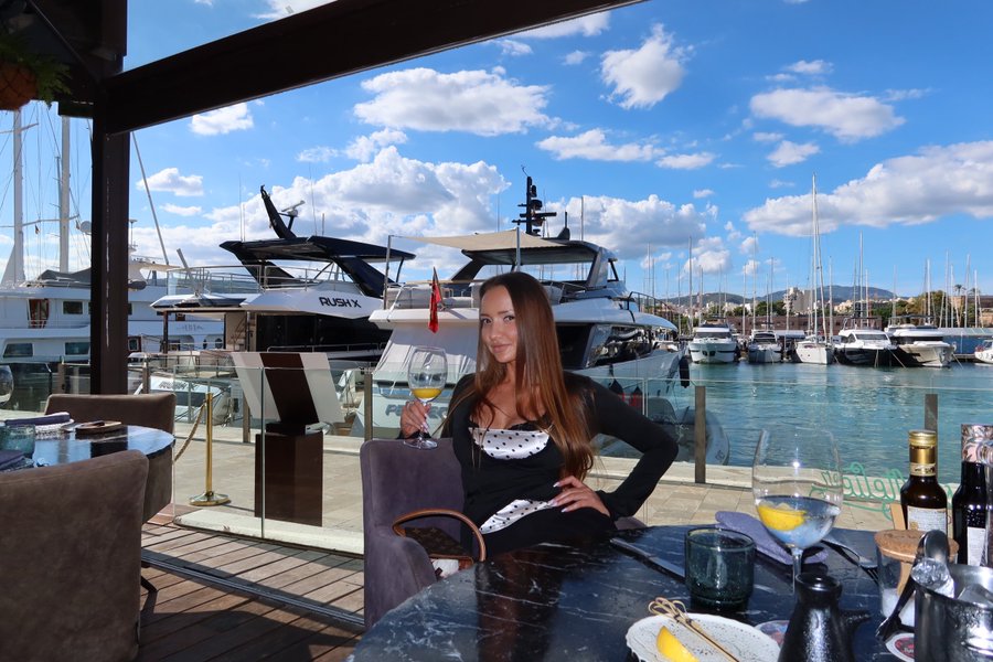 A woman in a polka-dot outfit holds a glass of white wine at a marina restaurant, with luxury yachts including one labeled "RUSHA" docked in the background.