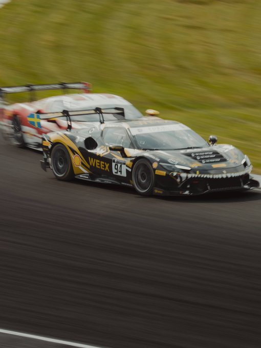 Two race cars speed along a track, with the leading black and gold car prominently displaying 'WEEX' and the number 94.